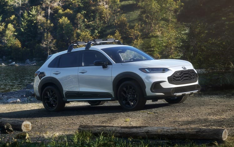 Vista de perfil desde el lado del pasajero de la Honda HR-V Sport 2025 en Platinum White Pearl con accesorios originales estacionada en una carretera boscosa.