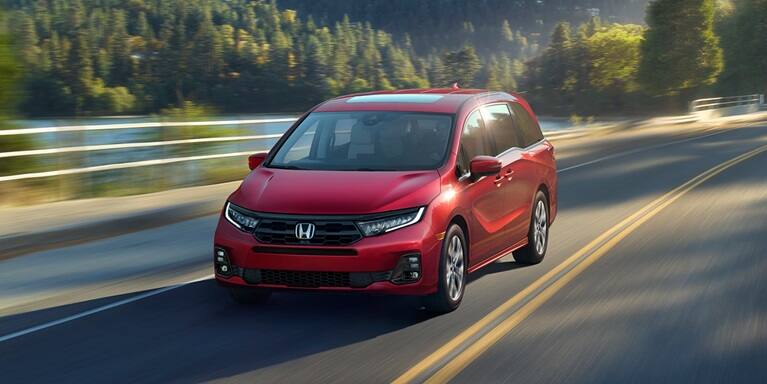 2027 Honda Odyssey Elite in Radiant Red Metallic II front driving on a scenic road near a lake and forest (Minivan).